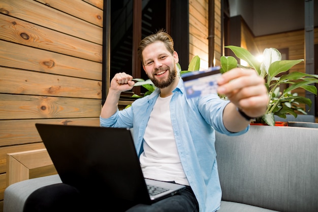 Uomo caucasico sorridente attraente che utilizza computer portatile nello spazio di lavoro congiunto, tenente la carta di credito