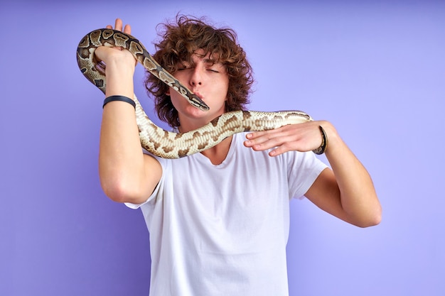 uomo che addestra un serpente e intrattiene, giovane ragazzo caucasico che gioca con lui, bacia un serpente, in posa isolato sul muro viola