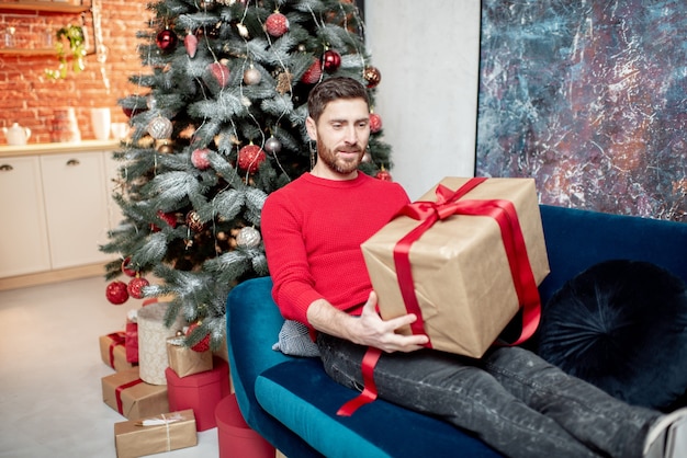 Uomo che apre una confezione regalo sdraiato sul divano durante le vacanze invernali con l'albero di Natale a casa