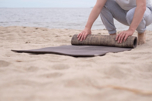 Uomo che arrotola il tappetino da yoga dopo l'allenamento sulla spiaggia sabbiosa