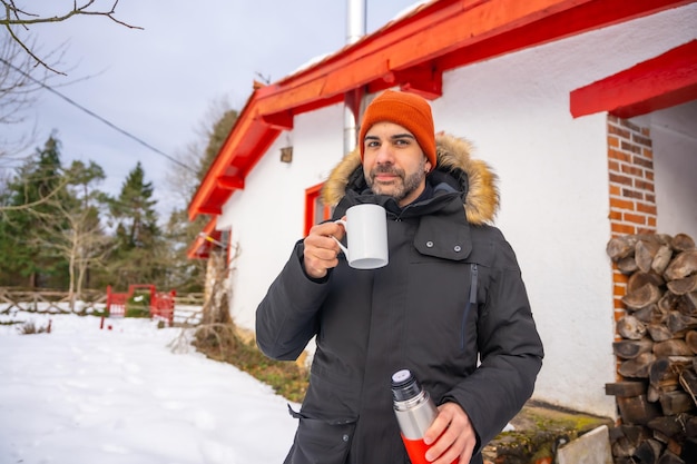 Uomo che beve caffè da un thermos caldo in inverno nella neve accanto al camino a legna