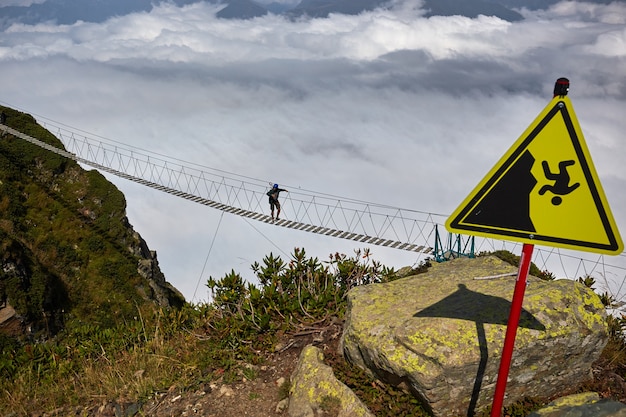 Uomo che cammina sul ponte sospeso e guarda le montagne nuvolose sottostanti.