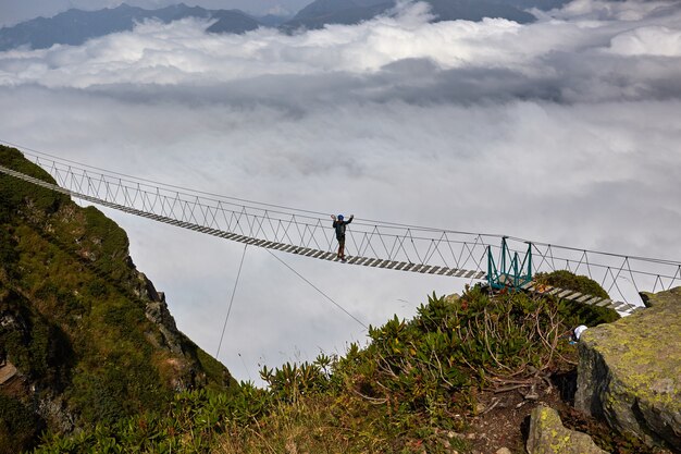 Uomo che cammina sul ponte sospeso e guarda le montagne nuvolose sottostanti.