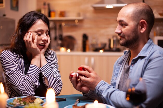 Uomo che celebra la relazione e chiede alla ragazza di sposarlo mentre cena. Uomo che chiede alla sua ragazza di sposarsi in cucina durante una cena romantica. Felice donna caucasica sorridente essendo discorso