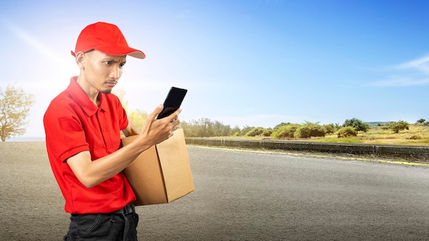 Uomo che consegna pacchi con un cappello che tiene un telefono cellulare mentre trasporta un pacco