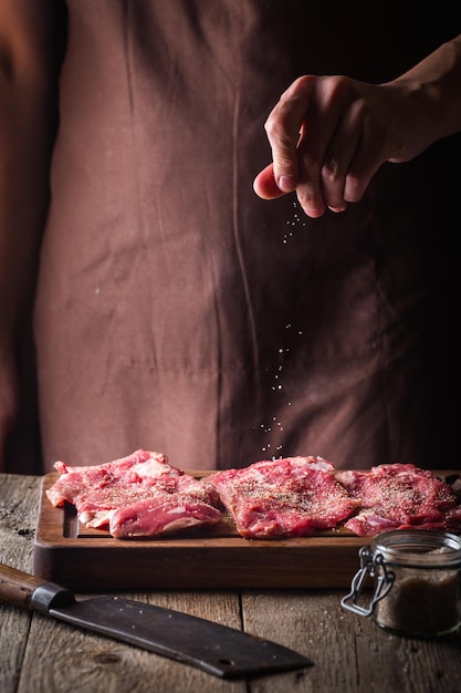 Uomo che cucina bistecche di carne in cucina. Lo chef sale e pepe su fondo di legno