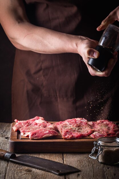 Uomo che cucina bistecche di carne in cucina. Lo chef sale e pepe su fondo di legno