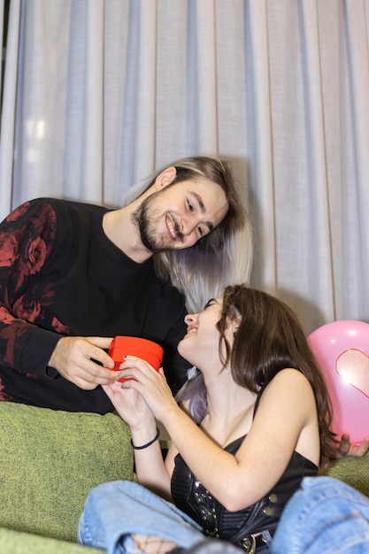 Uomo che dà un regalo alla sua ragazza seduta sul divano Ragazzo che dà un regalo di San Valentino alla sua ragazza seduta sul divano