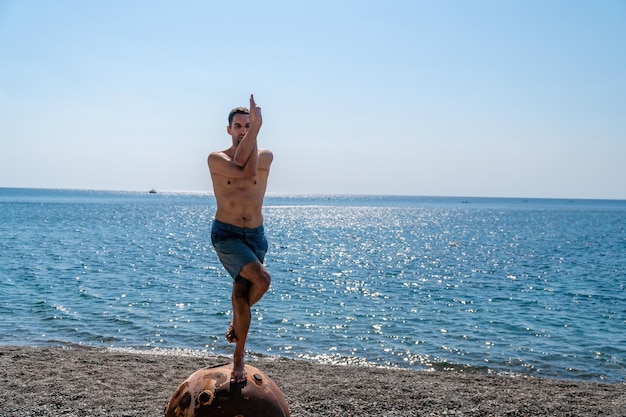 Uomo che fa esercizio di yoga all'aperto in piedi su una vecchia miniera marina galleggiante arrugginita sulla spiaggia con costa rocciosa e sfondo del mare Stile di vita sano inquinamento protezione della natura guerra e concetto di pace