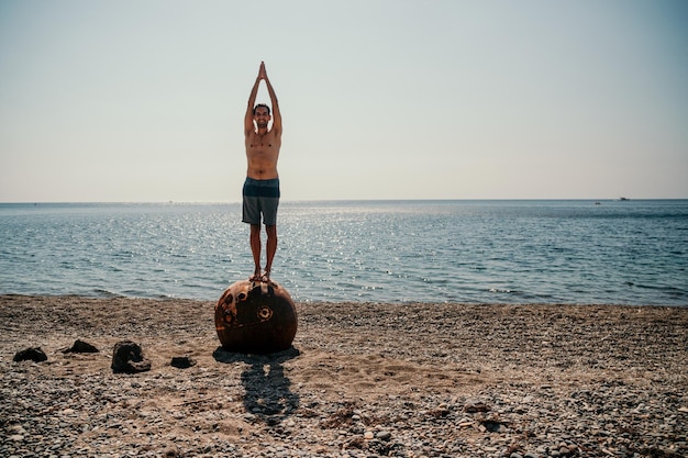Uomo che fa esercizio di yoga all'aperto in piedi su una vecchia miniera marina galleggiante arrugginita sulla spiaggia con rocce