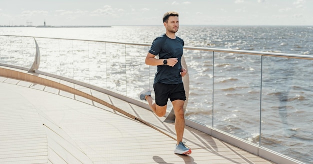 Uomo che fa jogging vicino al mare concentrato e in forma in abbigliamento sportivo con smartwatch e sfondo soleggiato