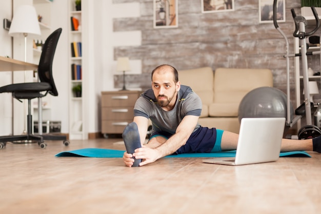 Uomo che fa le gambe che si allungano sul tappetino da yoga dall'allenamento online durante il blocco globale.