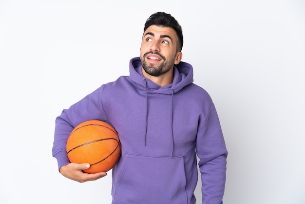 Uomo che gioca a basket sopra il muro bianco isolato pensando a un'idea mentre guarda in alto