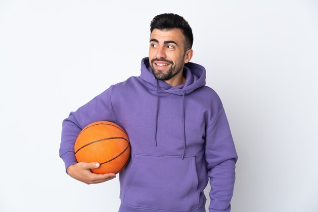 Uomo che gioca a basket sopra la parete bianca isolata che guarda al lato e che sorride