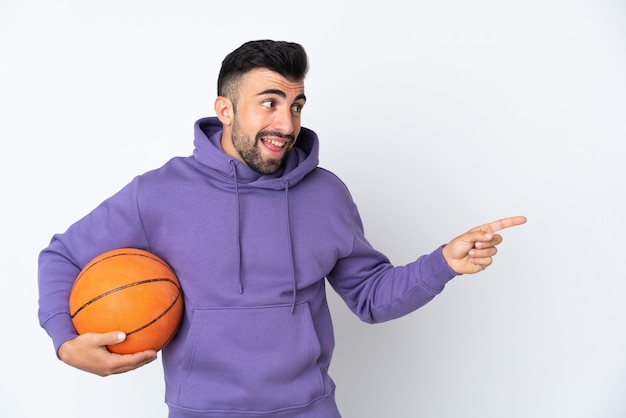 Uomo che gioca a basket sul muro bianco isolato che punta il dito di lato e presenta un prodotto