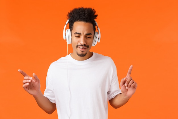 Uomo che gode di battiti fantastici. Ragazzo afroamericano attraente moderno hipster con taglio di capelli afro, baffi, occhi chiusi tamburellare con le dita e ascoltare musica in cuffia, arancione