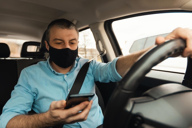 Uomo che guida l'auto utilizzando lo smartphone che indossa la maschera facciale in automobile