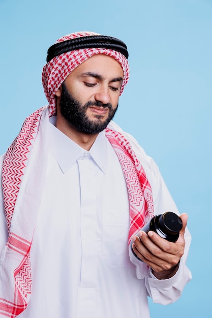 Uomo che indossa abiti musulmani tradizionali controlla le istruzioni sulla bottiglia di integratori con un'espressione infelice Arabo vestito in thobe e ghutra con farmaci per il trattamento delle malattie