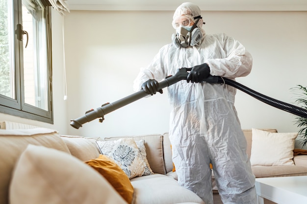 Uomo che indossa DPI disinfettando il soggiorno di una casa con una macchina disinfettante COVID-19. Pandemia del concetto di assistenza sanitaria