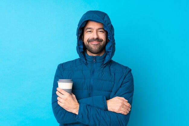 Uomo che indossa giacca invernale e in possesso di un caffè da asporto sul muro blu isolato mantenendo le braccia incrociate in posizione frontale