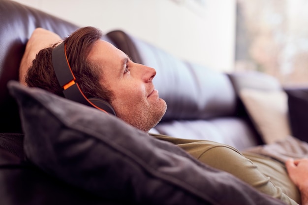 Uomo che indossa le cuffie senza fili che ascolta la musica sdraiato sul divano a casa indossando il pigiama