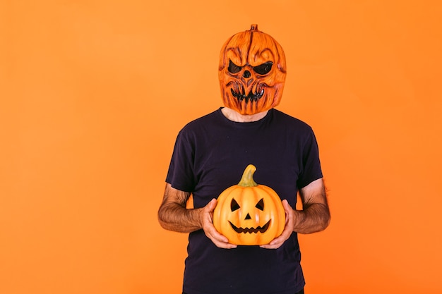 Uomo che indossa una spaventosa maschera in lattice di zucca con t-shirt blu, con in mano la zucca "Jack-o-lantern" su sfondo arancione. Halloween e giorni del concetto morto.