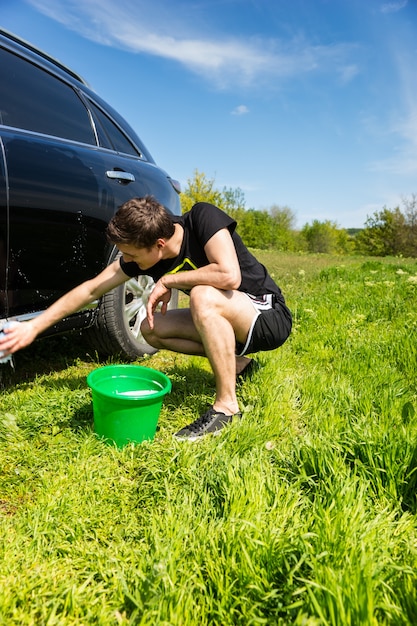 Uomo che lava auto con spugna insaponata, accovacciato accanto al secchio verde nel campo erboso verde in una luminosa giornata di sole con cielo blu