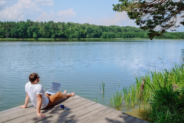 Uomo che lavora al computer portatile mentre è seduto su gambe di legno del bacino nell'acqua del fiume