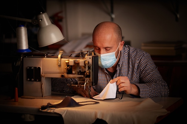 Uomo che lavora con la macchina da cucire facendo maschera facciale fatta in casa per prevenire e fermare la diffusione del virus corona