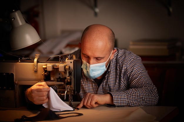 Uomo che lavora con la macchina da cucire facendo maschera facciale fatta in casa per prevenire e fermare la diffusione del virus corona