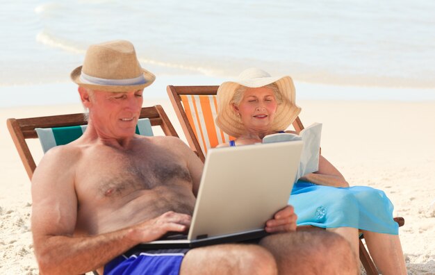 Uomo che lavora sul suo computer portatile mentre sua moglie sta leggendo in spiaggia