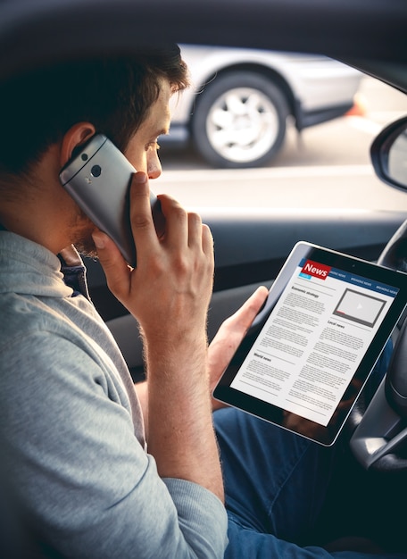 Uomo che legge notizie alla guida di un'auto con un tablet in mano parlando sul cellulare