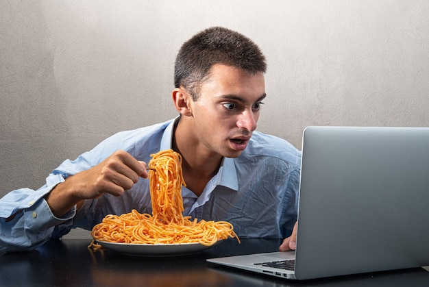 Uomo che mangia spaghetti con salsa di pomodoro e guarda il computer