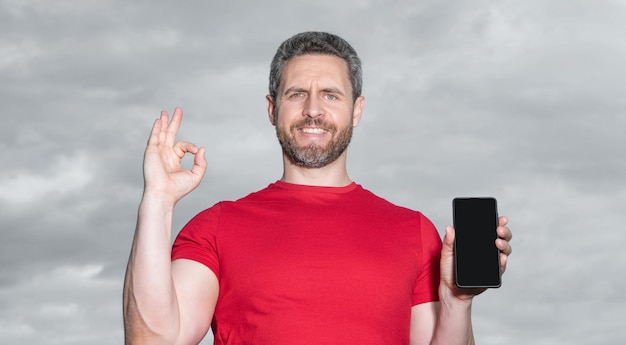 Uomo che mostra l'app del telefono sullo sfondo del cielo ok uomo che mostra lapp del telefono all'aperto uomo che mostra Lapp del cellulare indossa una maglietta rossa foto delluomo che mostra l‘app del telefono