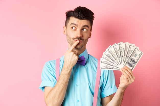 Uomo che pensa allo shopping, guardando pensieroso mentre si tiene denaro in dollari, in piedi sopra il rosa.