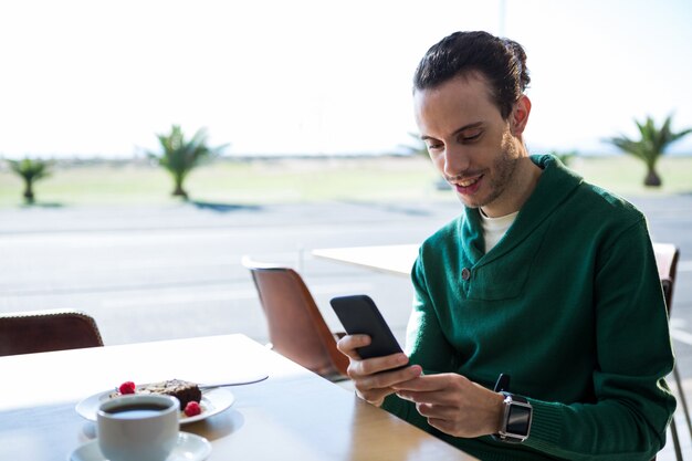 Uomo che per mezzo del telefono cellulare con il dolce e la tazza di caffè sulla tavola