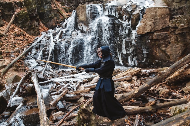 Uomo che pratica il kendo con la spada di bambù sulle rocce della cascata e sullo sfondo della foresta