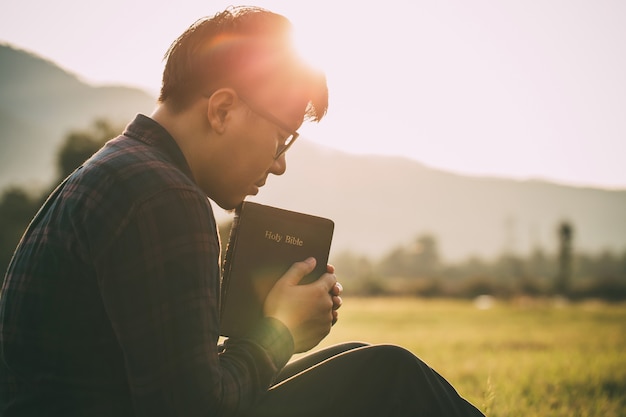 Uomo che prega sulla Sacra Bibbia in un campo durante un bellissimo tramonto maschio seduto con gli occhi chiusi
