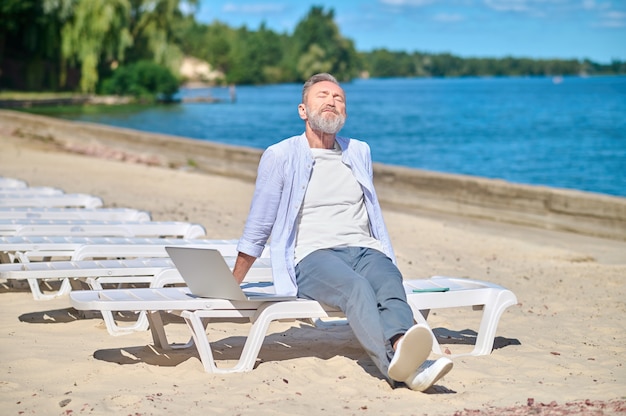 Uomo che prende il sole seduto con il computer portatile sulla spiaggia