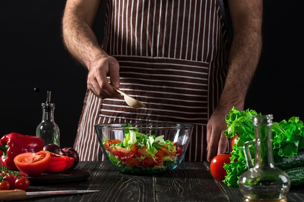 Uomo che prepara insalata con verdure fresche su un tavolo di legno. Cucinare cibi gustosi e sani. Su sfondo nero. Cibo vegetariano, concetto sano o di cucina.