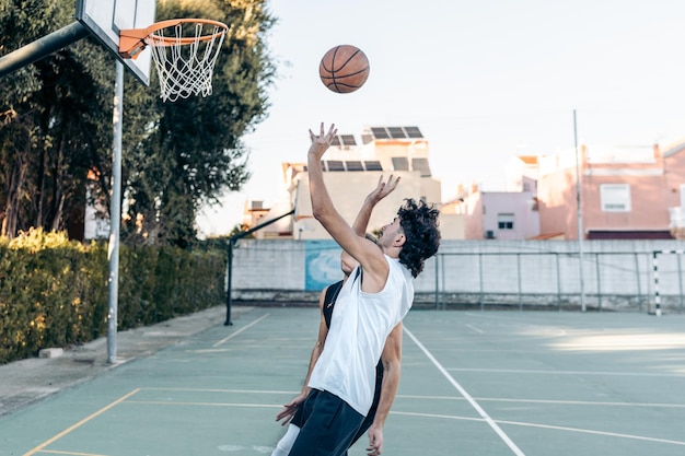 Uomo che salta mentre lancia una palla in un cesto giocando a basket in un campo urbano all'aperto