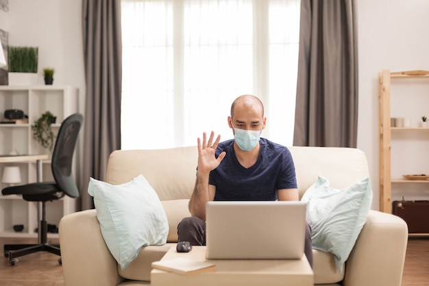 Uomo che saluta durante una videochiamata su laptop seduto o divano in tempo di pandemia globale.