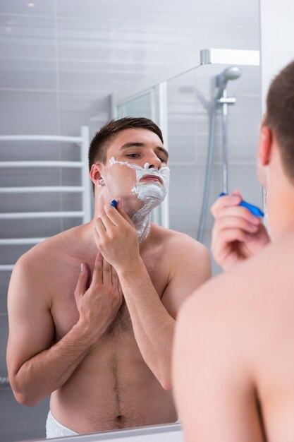 Uomo che si fa la barba con un rasoio e guarda lo specchio nel moderno bagno piastrellato di casa