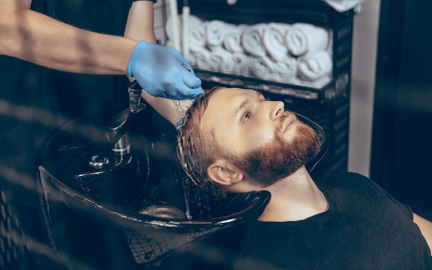 Uomo che si fa tagliare i capelli dal barbiere indossando la maschera durante la pandemia di coronavirus. Barbiere professionista che indossa guanti. Covid-19, concetto di bellezza, cura di sé, stile, assistenza sanitaria e medicina.