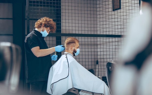 Uomo che si fa tagliare i capelli dal barbiere indossando la maschera durante la pandemia di coronavirus. Barbiere professionista che indossa guanti. Covid-19, concetto di bellezza, cura di sé, stile, assistenza sanitaria e medicina.