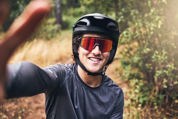 Uomo che si fa un selfie mentre si pedala su un sentiero naturalistico con indosso un casco e occhiali da sole Ritratto di un ciclista in bicicletta attraverso un parco o una foresta che scatta una foto sorridente e indossa l'equipaggiamento di sicurezza