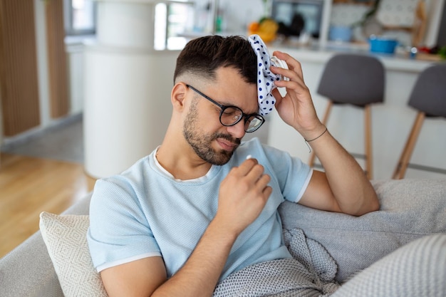 Uomo che si sente male seduto sul divano con mal di testa e applica la borsa del ghiaccio