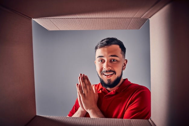 Uomo che sorride, disimballa e apre una confezione regalo di cartone e guarda dentro. Vista dalla scatola