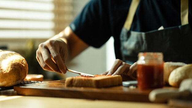 Uomo che spalma marmellata di lamponi su una fetta di pane con un coltello da tavola che prepara una sana colazione al tavolo della cucina