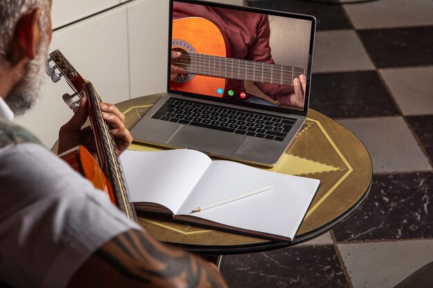 Uomo che suona la chitarra acustica e guarda le lezioni online sul laptop mentre si esercita a casa... Formazione online, lezioni online.